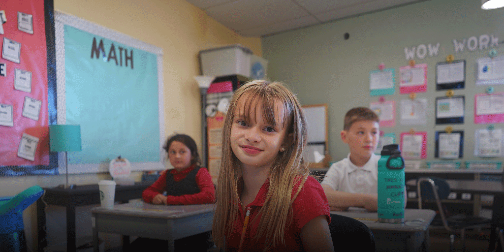 Smiling student at a desk in a classroom with a math bulletin board.