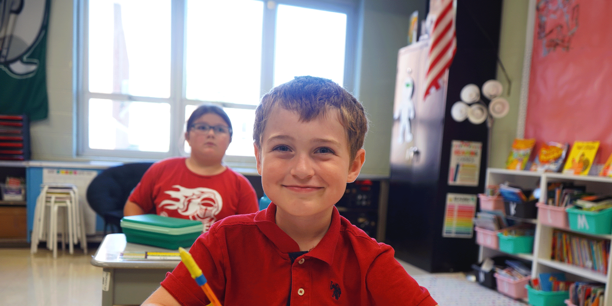 Smiling student in a red shirt at a classroom desk.