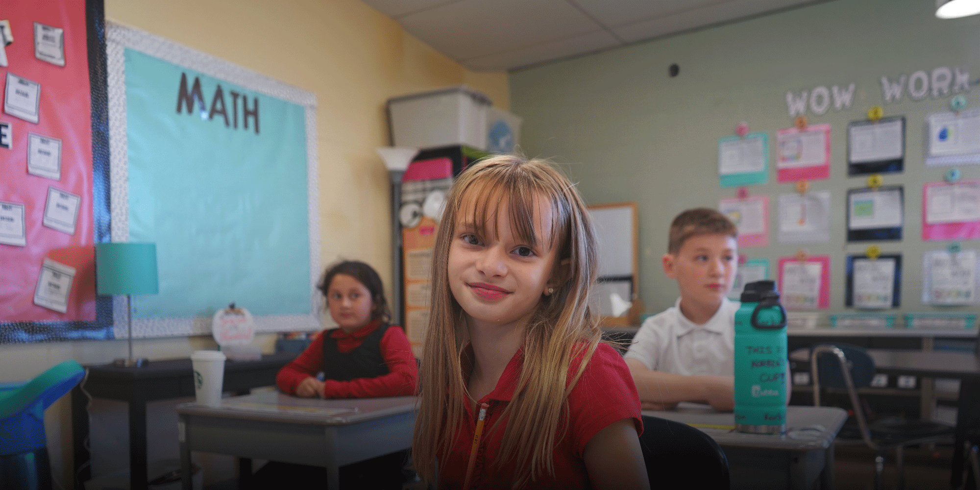 Smiling student at a desk in a classroom with a math bulletin board.